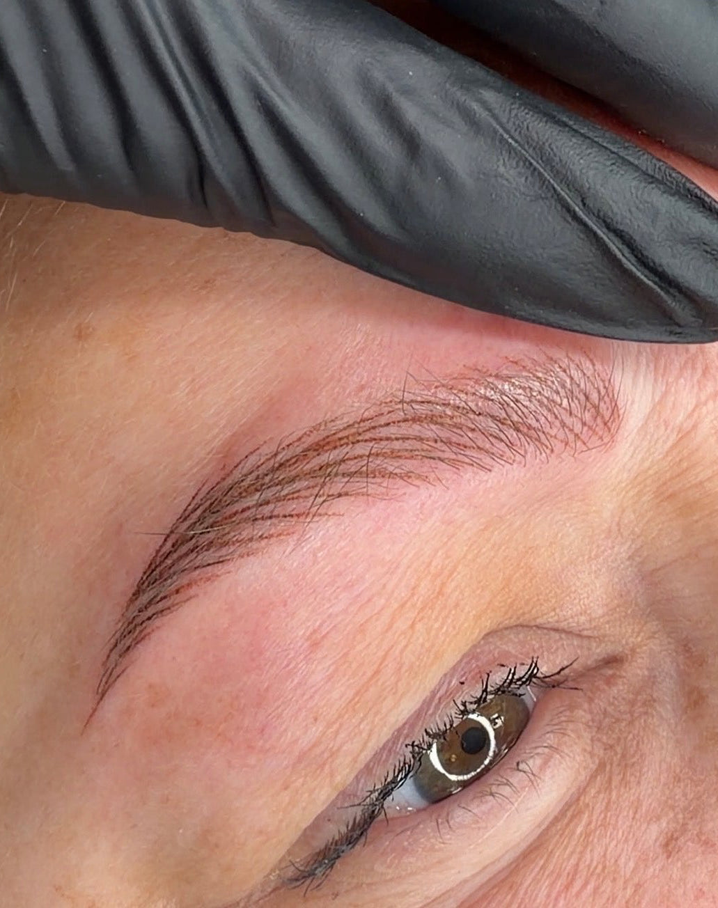 Close-up of a person's face with a freshly shaped eyebrow, showing the eyebrow, eye, skin, and a black gloved hand holding the skin in place.